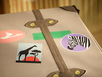 Close-up of a storage box or chest covered in gray fabric, with decorative brown straps and travel-style stickers featuring a zebra, a giraffe, and a flamingo.