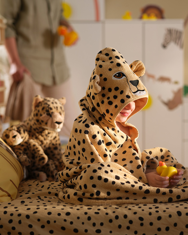 Un niño con una toalla SANDLÖPARE con capucha y estampado de guepardo está sentado en una cama rodeado de peluches de guepardo y un patito de goma.