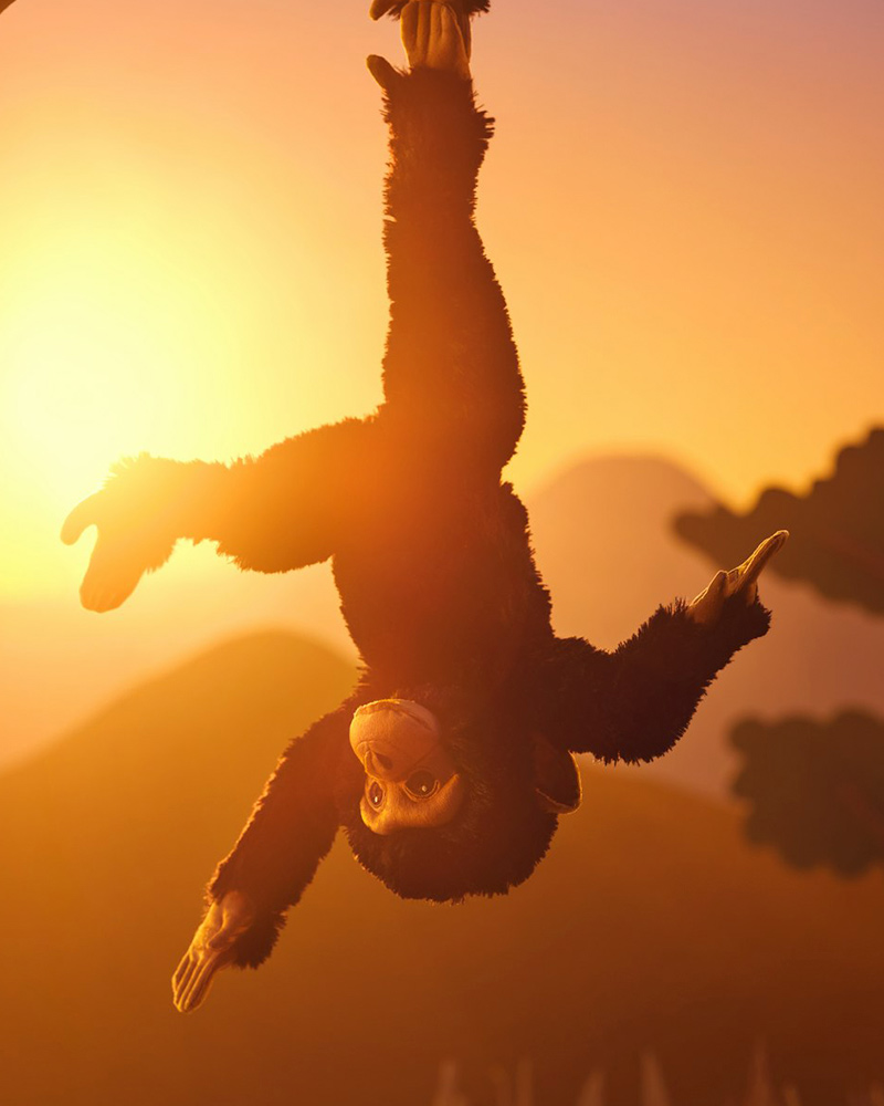 Un chimpancé de peluche negro de la colección SANDLÖPARE cuelga de sus pies en un paisaje de sabana soleado.