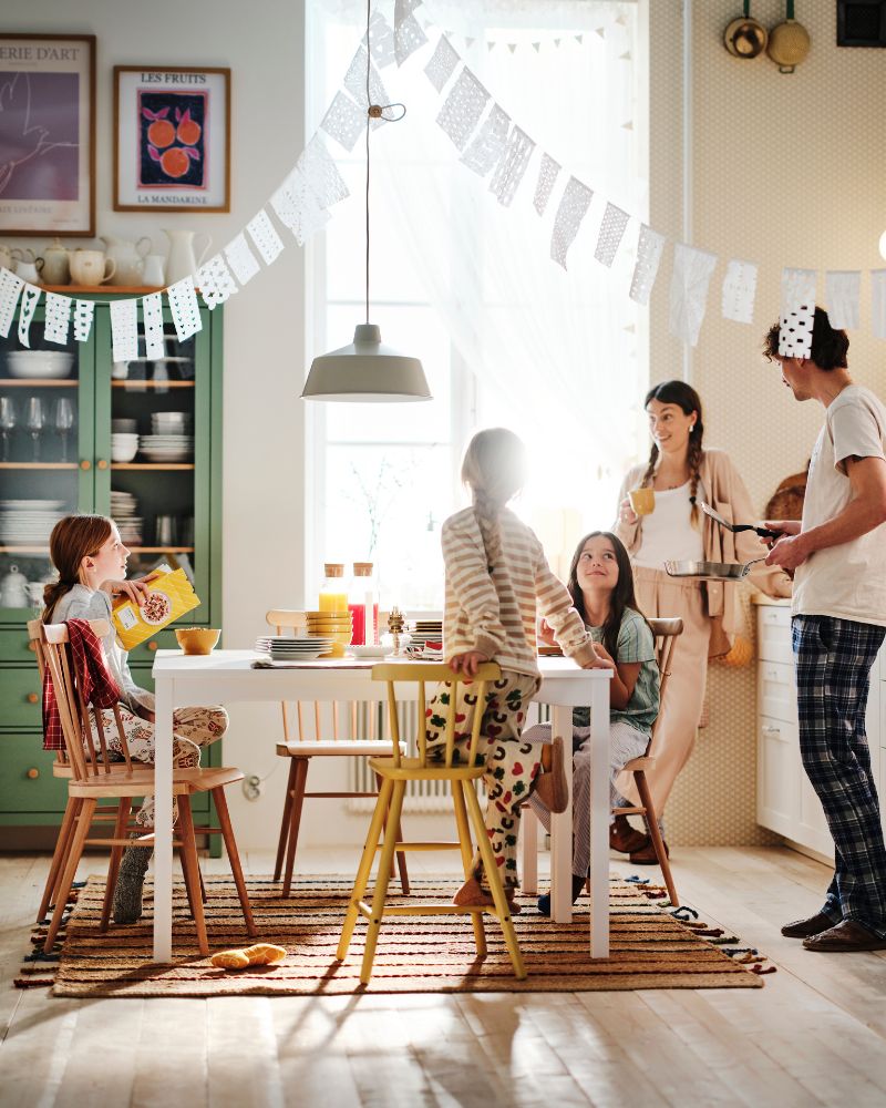 Una madre y un padre están de pie en la cocina mirando la mesa donde sus tres hijos están sentados disfrutando del desayuno.