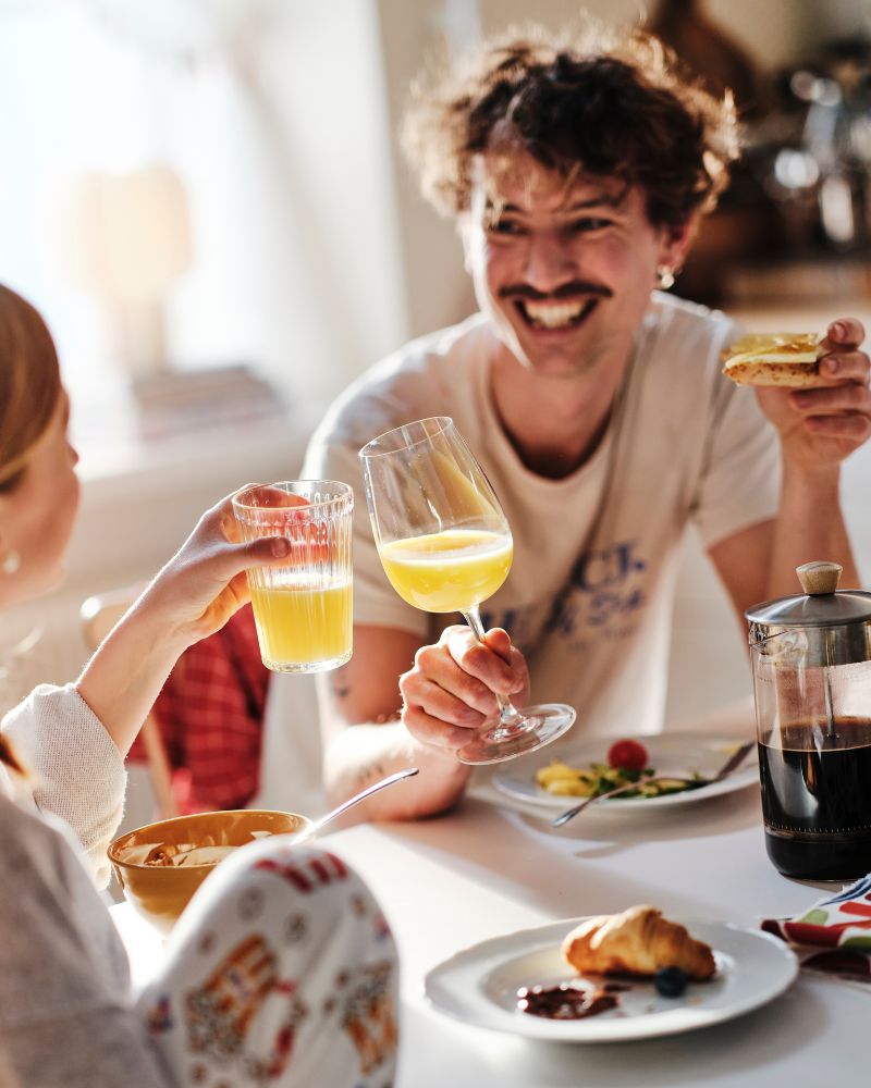Un padre sonriente y su hija adolescente, sentados a la mesa del desayuno, chocan sus vasos de zumo de naranja.