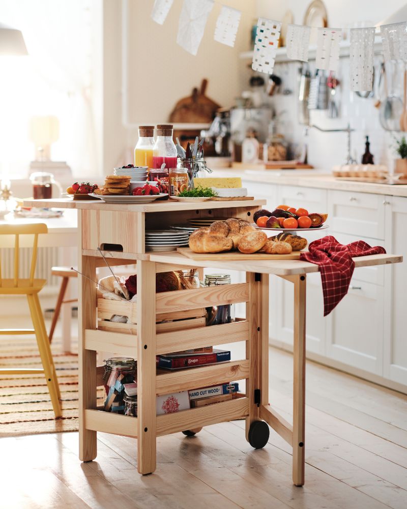 Mesa plegable móvil RESARÖ llena de comida para el desayuno y vajilla, colocada junto a una mesa de comedor en una cocina luminosa.