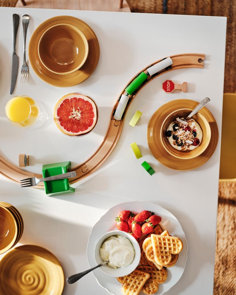 Plano superior de una mesa de desayuno con un plato de waes y varios cuencos. Una vía de tren de juguete serpentea entre ellos.
