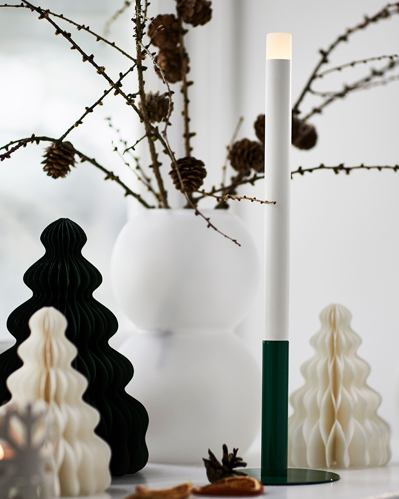 Una vela LED STRÅLA con base verde frente a adornos decorativos sobre una mesa iluminada.