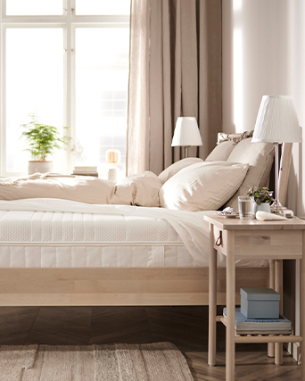 Dormitorio luminoso con cama de madera natural, ropa de cama beige y mesita de noche con lámpara. Líneas limpias y tonos neutros cálidos crean un espacio de descanso tranquilo