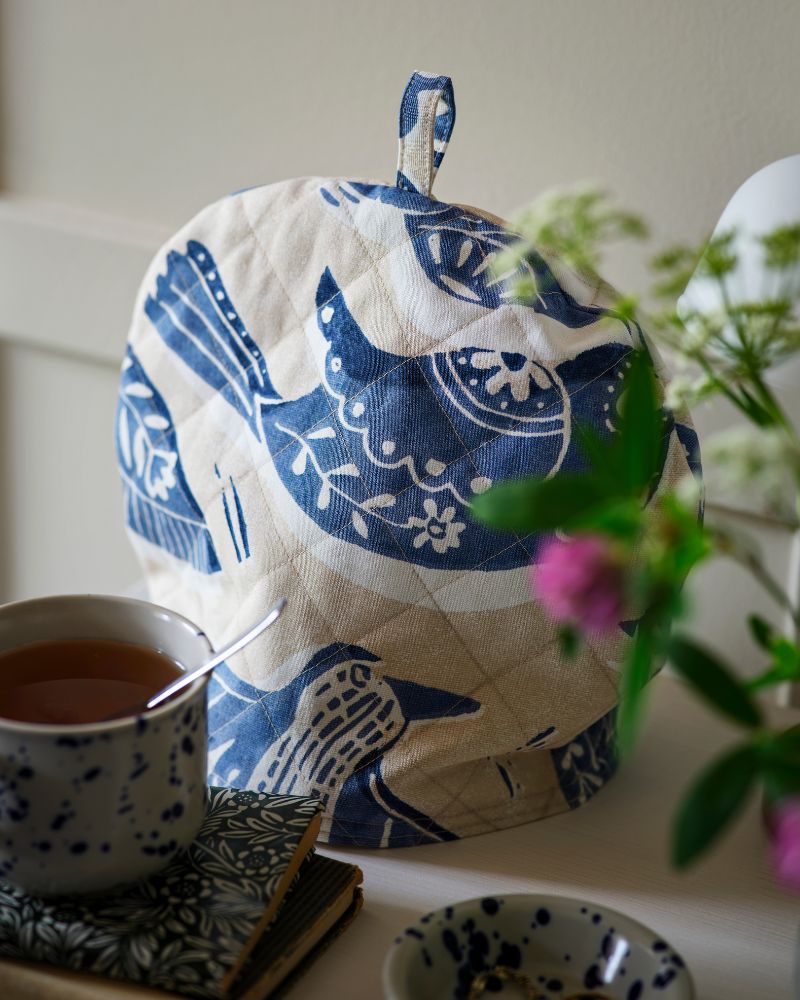 Un cubreteiera elaborado con tela precortada BITTERSÖTA en tonos beige y azul se encuentra sobre una tetera, junto a una taza y algunos cuadernos.