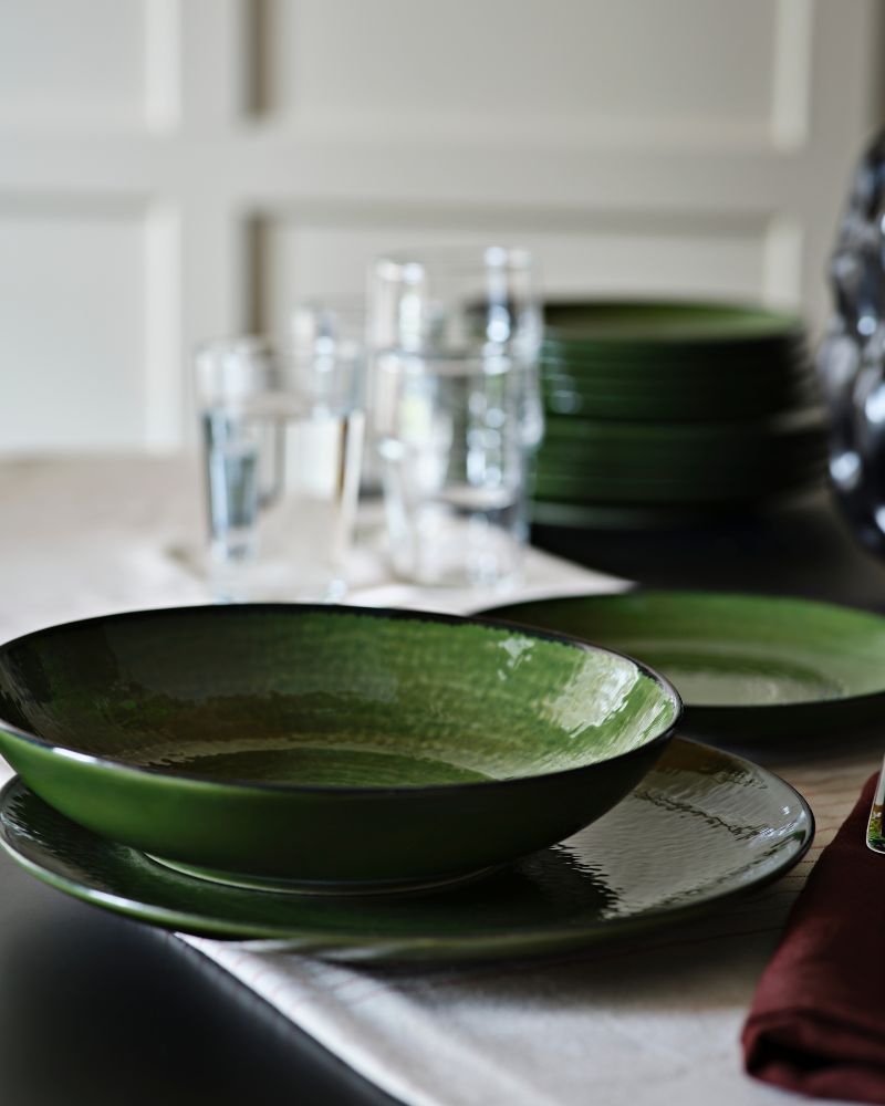 Un plato hondo PELARKAKTUS verde se encuentra sobre una mesa junto a cristalería y platos apilados en un ambiente de comedor elegante.