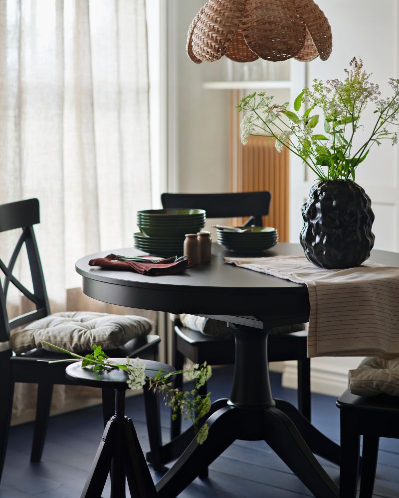 Una mesa extensible ROSENTORP negra está dispuesta con sillas, vajilla y un jarrón con flores en un comedor lleno de luz.
