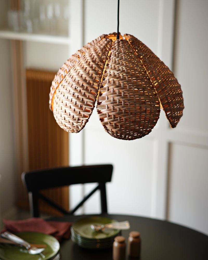 Una pantalla de lámpara colgante SANDKORN de tonos cálidos, con un diseño tejido, colgando sobre una mesa de comedor con platos verdes.