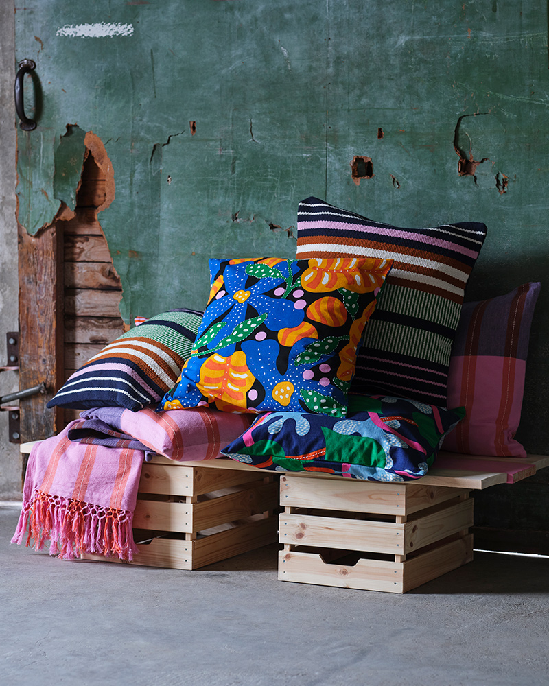 Sobre un par de cajas de madera hay fundas de cojín y mantas en rosa, azul, verde y naranja de la colección MÄVINN.