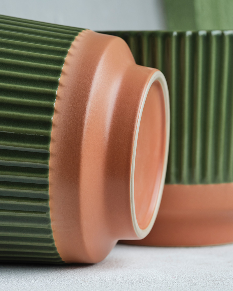 Una maceta verde de la colección MÄVINN, hecha de gres con relieves y base de terracota, yace de lado. Detrás hay otra maceta.