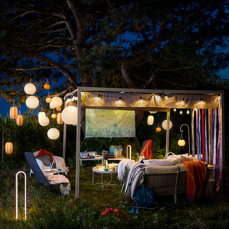 Un espacio exterior por la noche con lámparas colgantes SOLVINDEN de color naranja y blanco a rayas, alimentadas con energía solar, que cuelgan de un gran árbol.