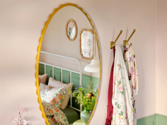 Un espejo DAGGKAPRIFOL de color amarillo oscuro refleja un acogedor dormitorio con decoración floral y una cama adornada con colores suaves.