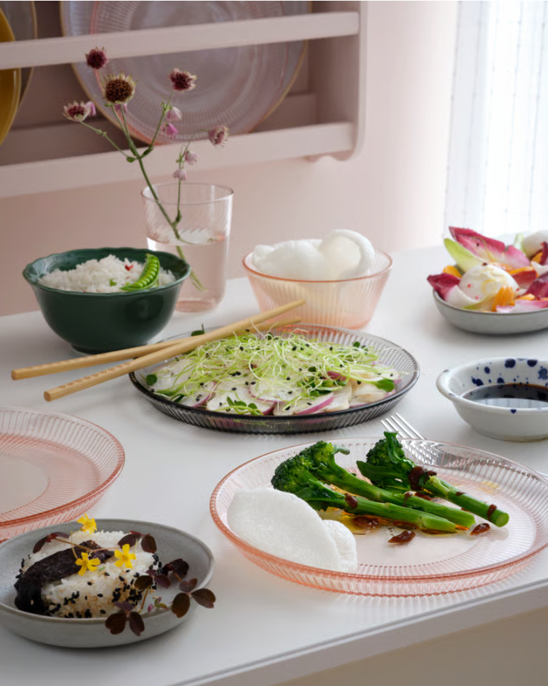 Un plato llano HAVSGÄDDA rosa se muestra con elegancia con varios platos, incluyendo ensalada, arroz y verduras.