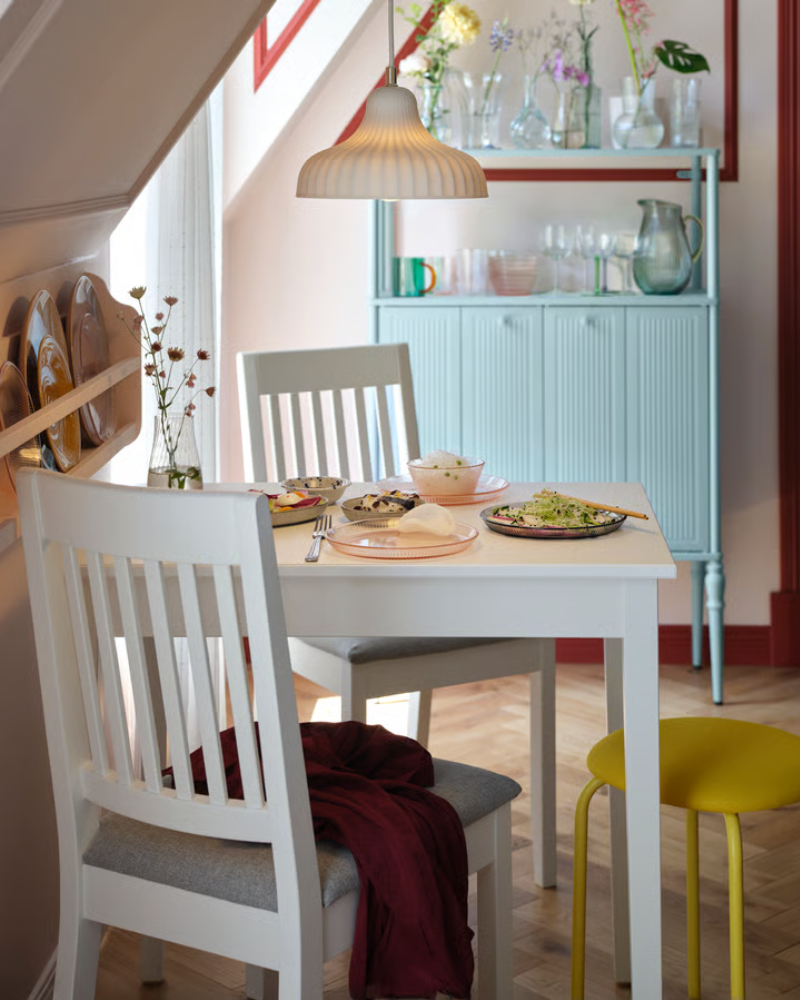 Una mesa NÄSINGE blanca se alza elegante en un luminoso comedor con un armario SÅGMÄSTARE gris azulado claro al fondo.