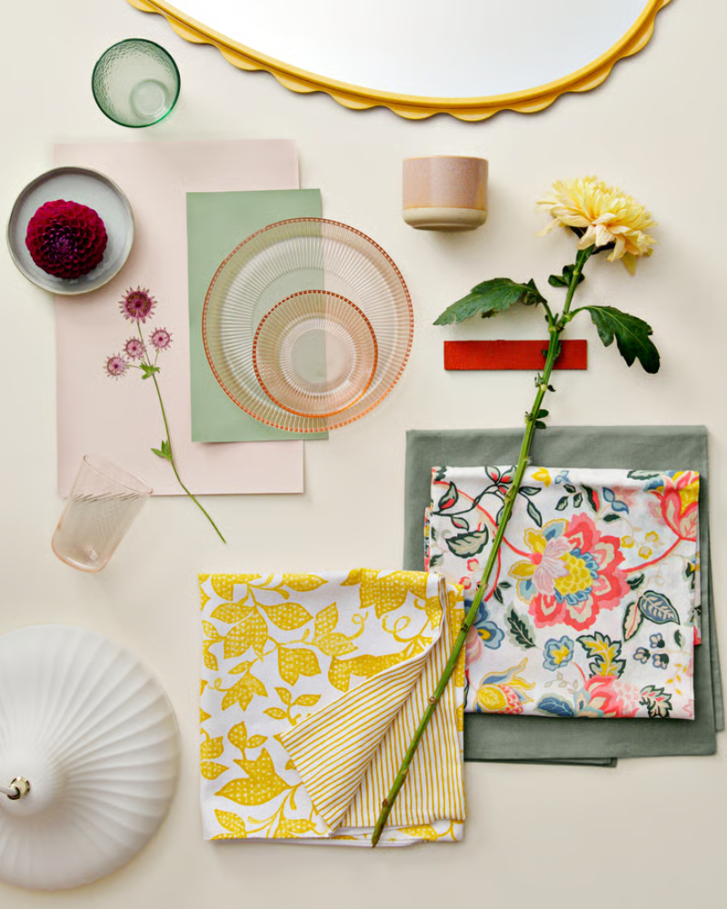 Una funda de cojín FJUNKLOCKA plegada en amarillo y blanco colocada entre otros artículos, flores y textiles en una elegante composición.