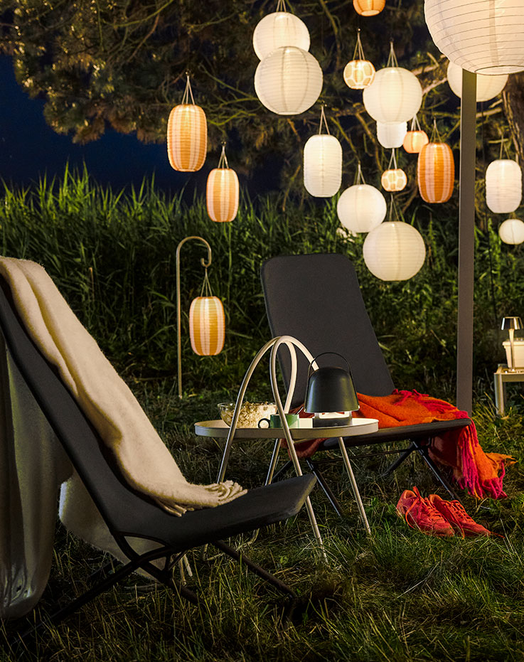 Una acogedora zona ajardinada para la noche con luces colgantes cuenta con un sillón VIGGSÖ gris oscuro, rodeado de vegetación.