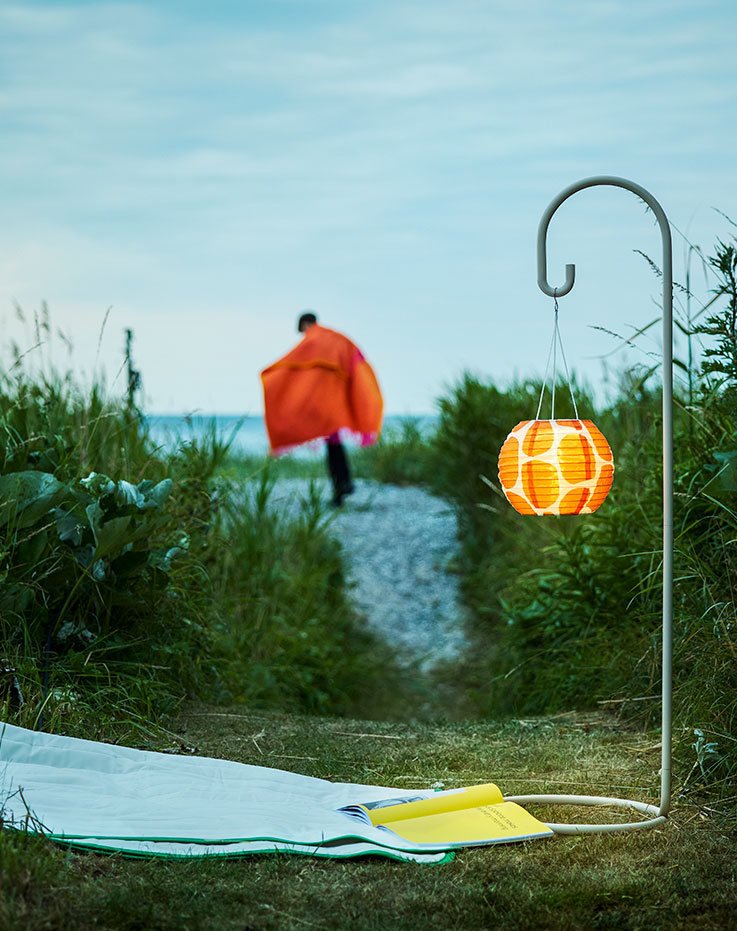 Una escena nocturna en un jardín muestra una lámpara SOLVINDEN de color beige con una pantalla naranja iluminada y una persona caminando.