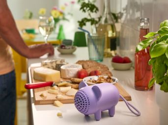 A purple mouse-shaped GREJSIMOJS portable Bluetooth speaker is on a kitchen worktop. A charcuterie board is behind it.