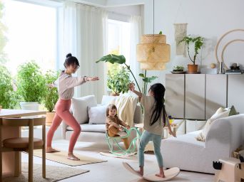 In a living room, a parent practices yoga as their children play on a GREJSIMOJS balance board and GREJSIMOJS rocking-bird.