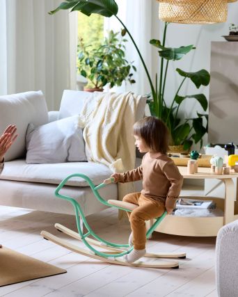 In a light and plant-filled living room, a parent on a yoga mat watches as their child climbs on a GREJSIMOJS moose stool.
