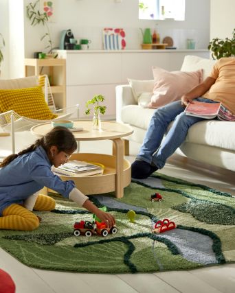 In a sunlit living room, a child sits on a GREJSIMOJS rug and plays with a train set while their parent sits on the sofa.