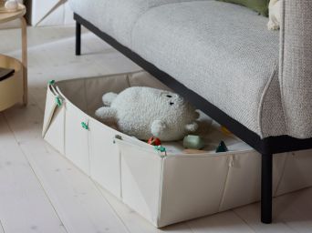 A beige GREJSIMOJS mat/foldable storage box pokes out from under a sofa in a modern living room. Soft toys are in the box.