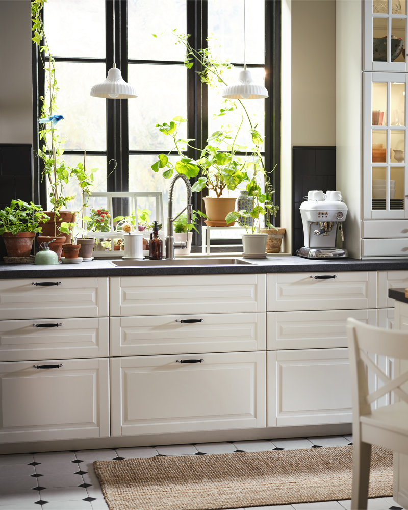 Una cocina tradicional con frentes de cocina BODBYN en color blanco roto, grandes ventanas con plantas y una cafetera.