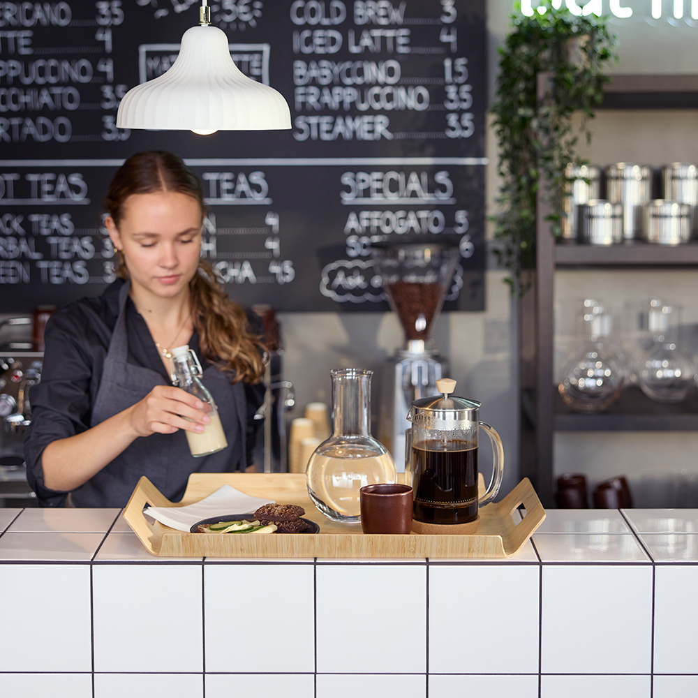 Café scene with a barista preparing a bamboo SILVERHAJMAL tray to serve drinks and food on a tiled café counter.