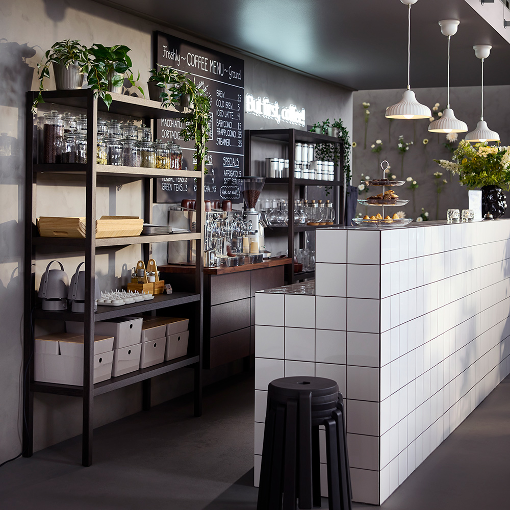A café counter with dark brown STOCKHOLM 2025 shelving units in oak veneer/dark brown filled with glassware and jars.