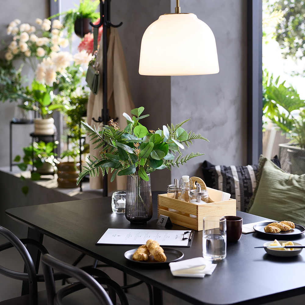 Close-up of a café with a black LISABO table, served food, a grey vase with greenery and flowers in the background.