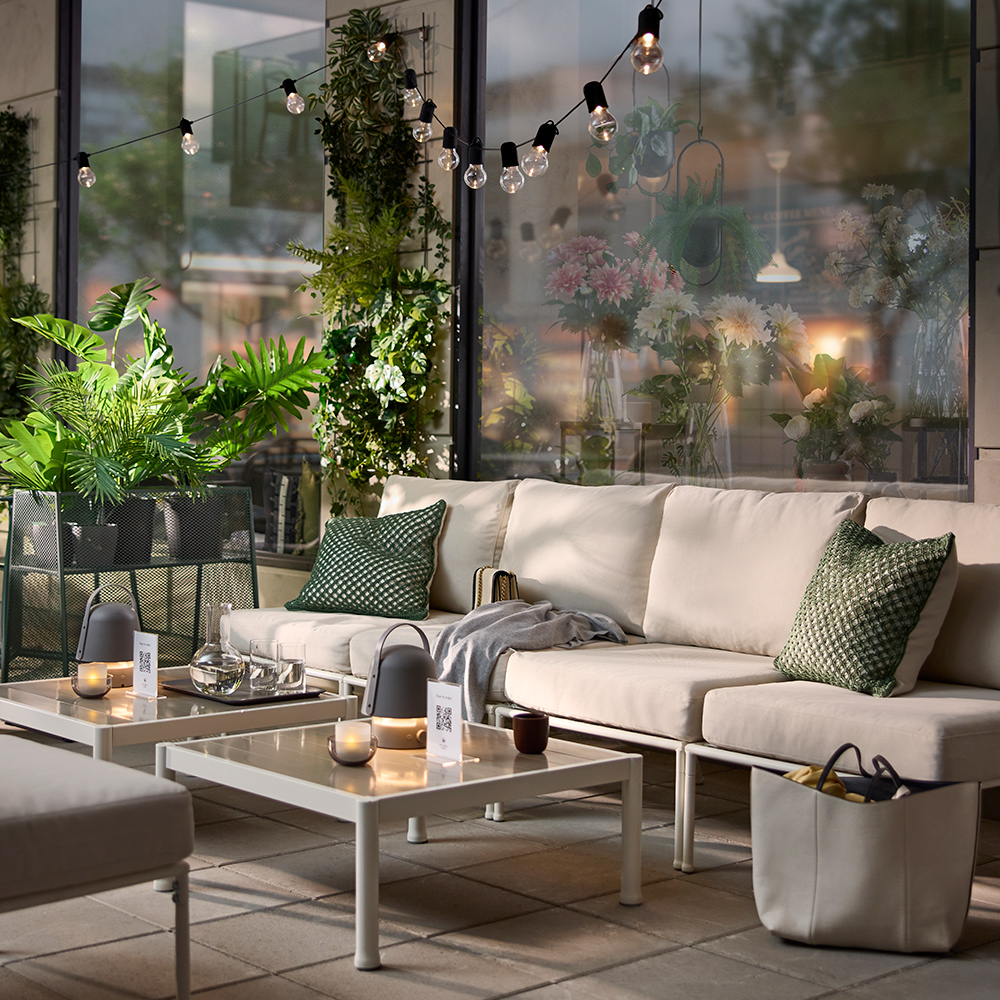 An outdoor café area with a white/beige SEGERÖN seat section for a modular sofa, surrounded by green plants and cosy lights.