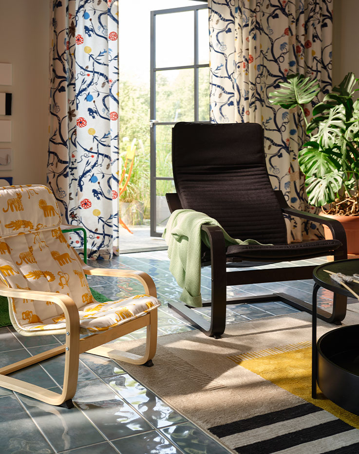 Un sillón POÄNG marrón oscuro junto a un sillón POÄNG más pequeño con estampado animal en una habitación con plantas y cortinas estampadas.