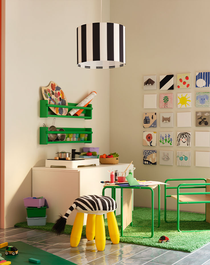 Una esquina con una silla y mesa infantil SMÅSKRAKE de color verde junto a una cocina de juguete STENMUS y dibujos de colores.