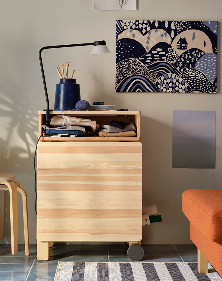 Una sala de estar soleada con una mesa plegable RESARÖ de pino en modo plegado, junto a una pared y al lado de unos taburetes KYRRE apilados