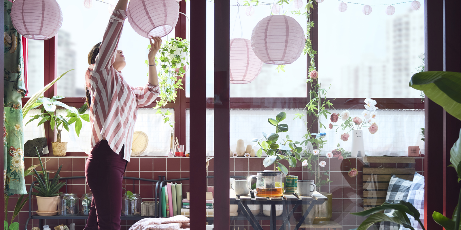 Un balcón cerrado con una mesa de exterior TÄRNÖ en negro y marrón claro, rodeada de plantas y lámparas decorativas de color rosa.
