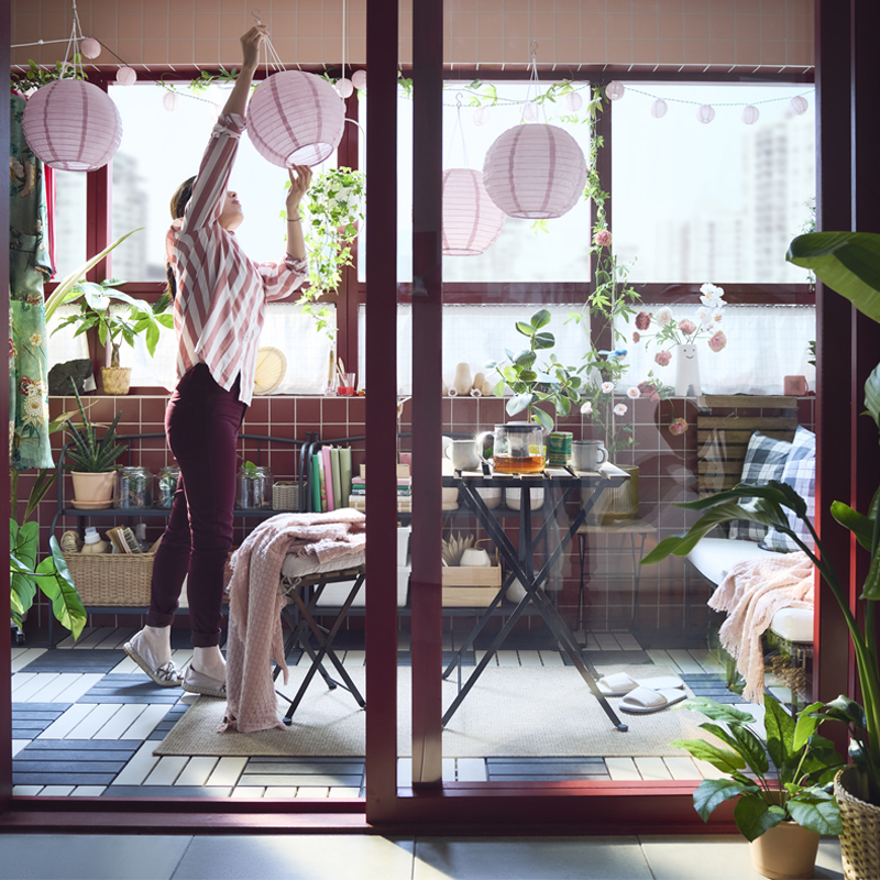 Un balcón cerrado con una mesa de exterior TÄRNÖ en negro y marrón claro, rodeada de plantas y lámparas decorativas de color rosa.