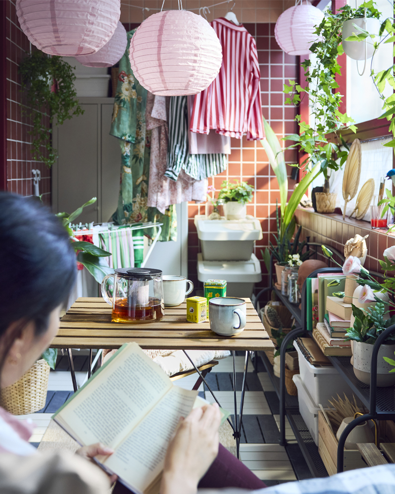 Un balcón alegre con plantas verdes y lámparas rosas, con una mesa de exterior TÄRNÖ de madera negra y marrón claro y tazas de té.