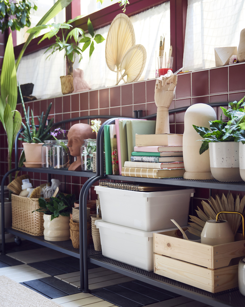 Un luminoso balcón cerrado con dos estanterías LÄCKÖ grises diseñadas para uso en exteriores, en las que se exponen diversas plantas y libros.