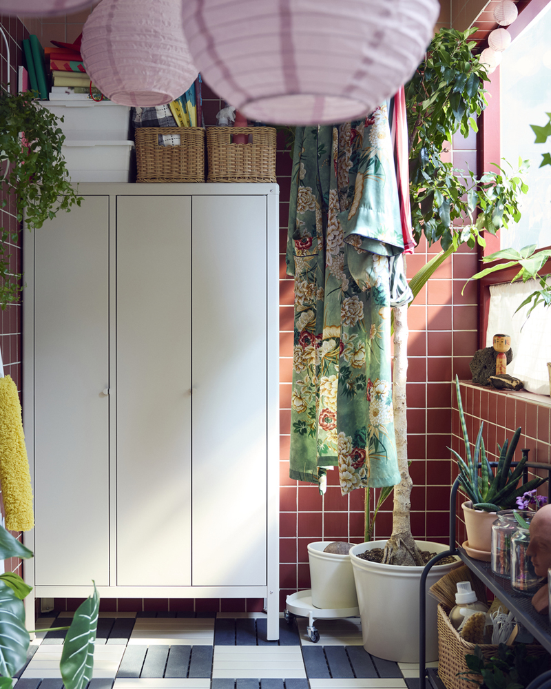 Un balcón con un armario KOLBJÖRN beige diseñado para uso en exteriores, rodeado de plantas colgantes, ropa y lámparas rosas.