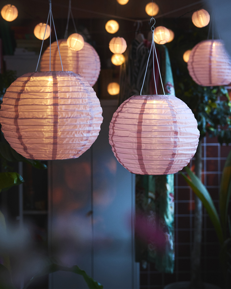 Un balcón decorado con exuberantes plantas cuenta con varias lámparas colgantes SOLVINDEN LED de energía solar de color rosa que cuelgan en la parte superior.