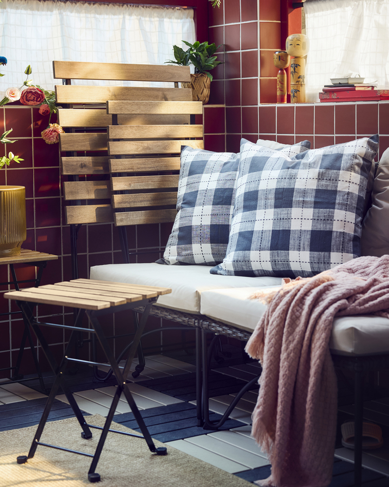 Un balcón con un sillón JUTHOLMEN gris oscuro y cojines gris claro-beige rodeados de almohadas estampadas y plantas verdes.