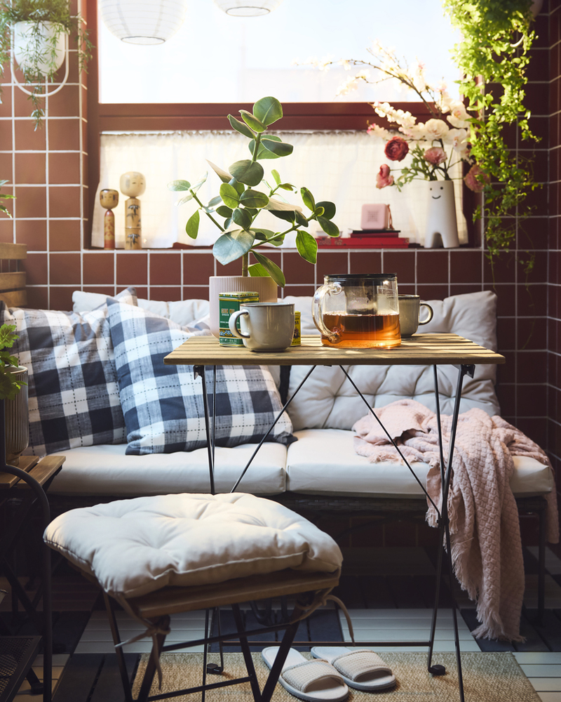 Un acogedor balcón con una mesa de exterior TÄRNÖ negra, rodeada de plantas, cojines y una jarra de cristal con té.
