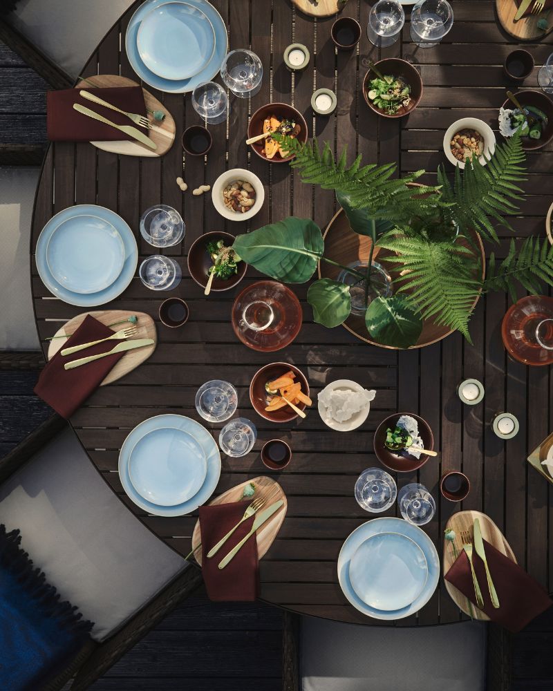 Una mesa de comedor al aire libre con una mesa redonda VITTSKÄR gris oscuro decorada con vajilla FÄRGKLAR azul claro y plantas.