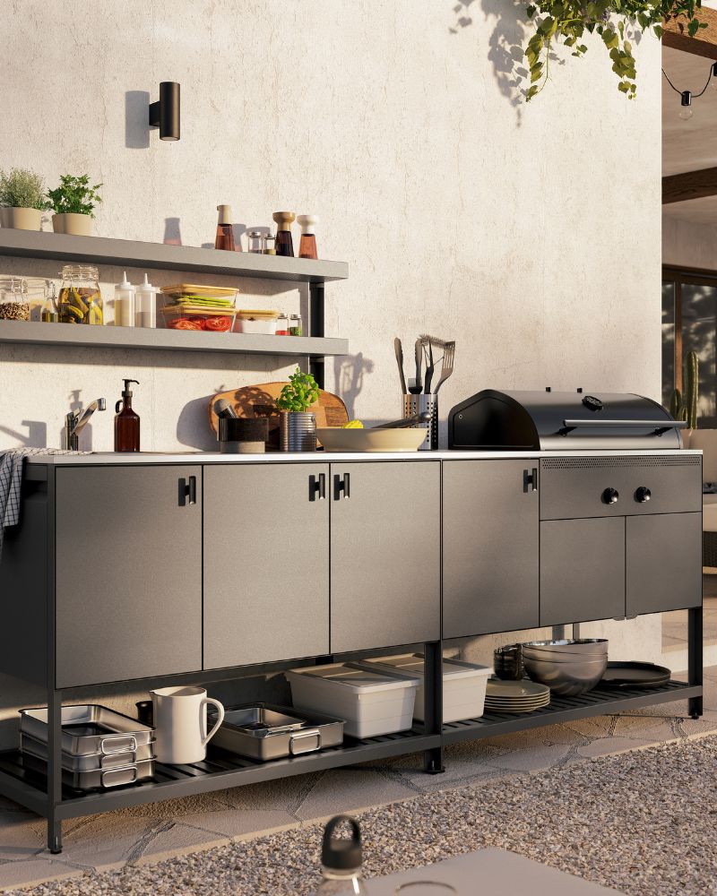 Una cocina exterior BÅTSKÄR de color gris oscuro con barbacoa de carbón. Sobre los armarios cerrados hay dos estantes abiertos con hierbas frescas.
