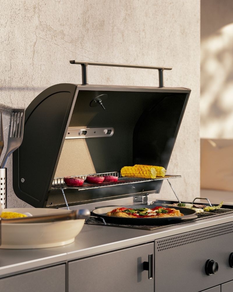 Un primer plano de la zona de barbacoa de una cocina exterior BÅTSKÄR de color gris oscuro. La tapa está abierta, dejando ver una pizza y verduras.