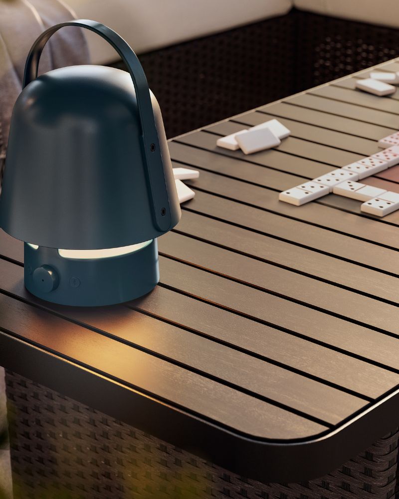 Una lámpara con altavoz Bluetooth VAPPEBY azul oscuro para uso en exteriores sobre una mesa de centro VITTSKÄR gris oscuro. Luz de la tarde al aire libre.