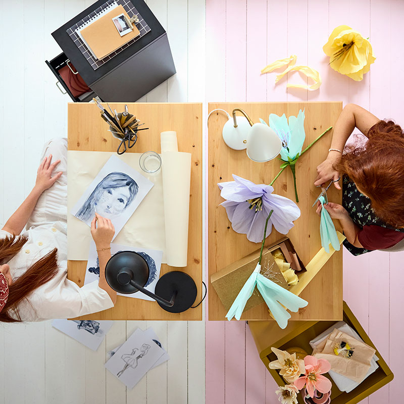 Dos escritorios BOLLPOJKE en pino/blanco uno al lado del otro, vistos desde arriba, con dos adolescentes haciendo manualidades y usando materiales de arte.