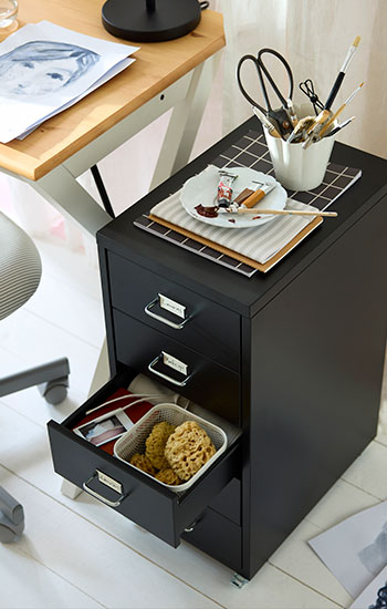 Cajonera HELMER negra con ruedas junto a un escritorio BOLLPOJKE, con material artístico y un cajón abierto.