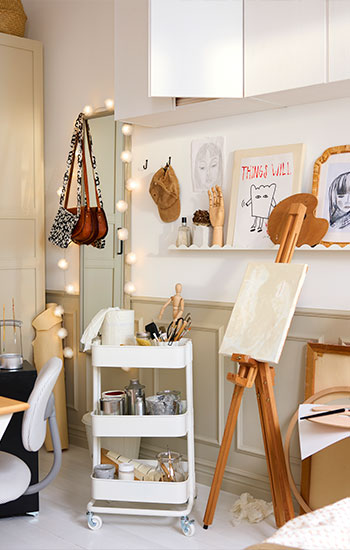 Un área de manualidades creativas con un carrito RÅSKOG blanco que contiene materiales de arte junto a un escritorio y un caballete en una habitación luminosa.